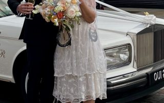 Bride and groom celebrating with champagne beside a classic white Rolls-Royce wedding car