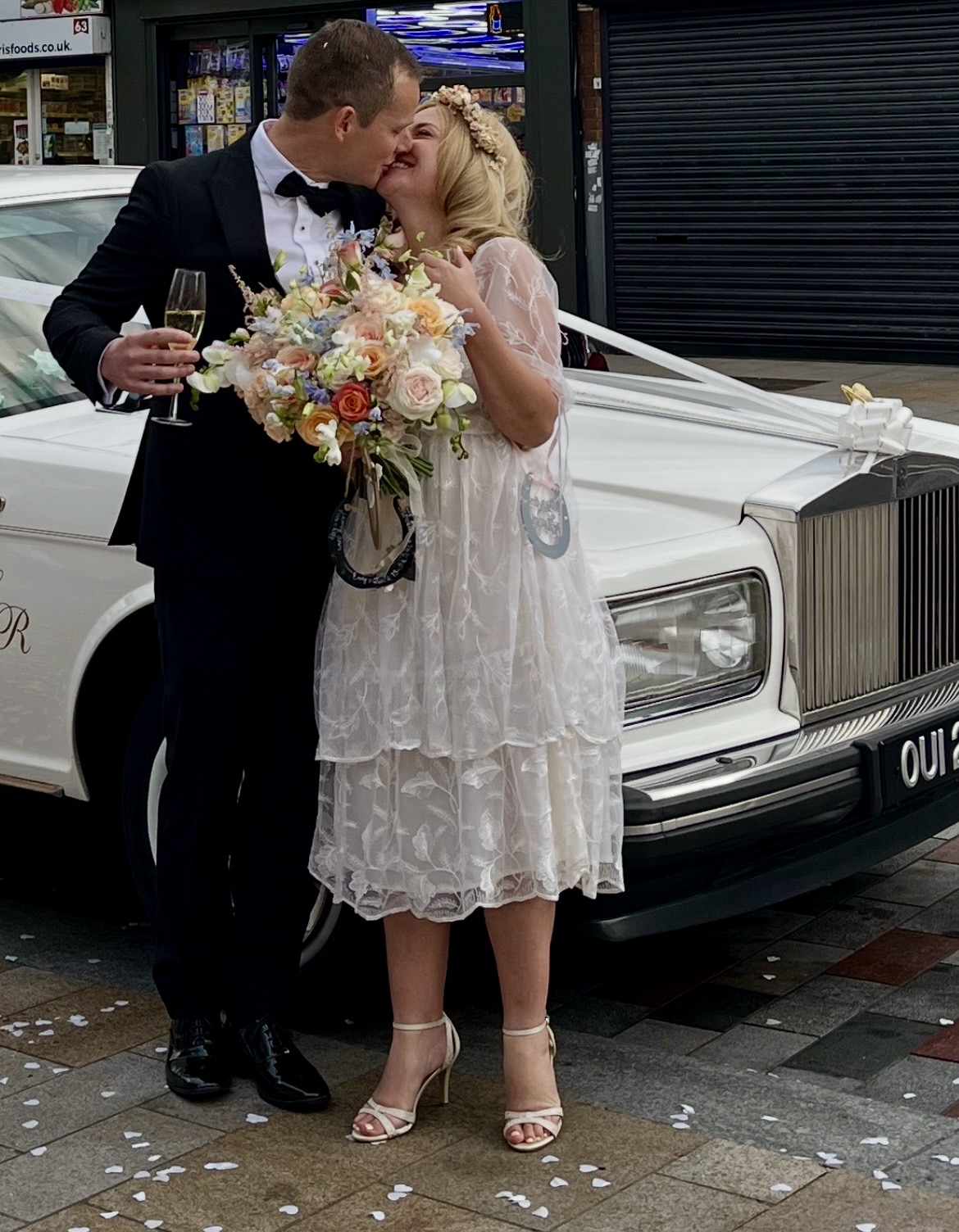 Bride and groom celebrating with champagne beside a classic white Rolls-Royce wedding car